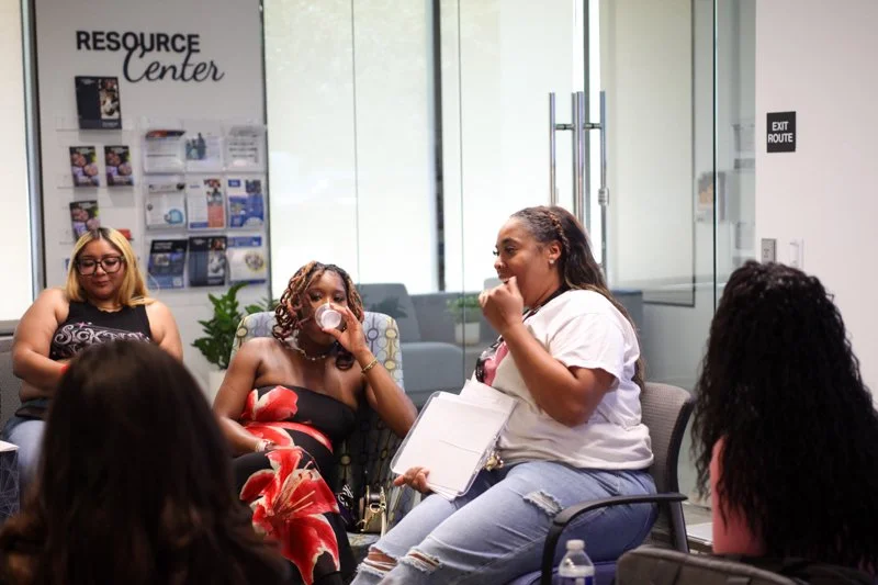 Six women seated in a glass-walled room, engaged in a discussion. One woman is drinking from a cup, another holds a notepad, and the others listen. The room has a Resource Center poster on the wall.