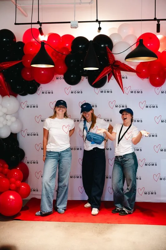 Three women standing on a red carpet in front of a backdrop with hearts and the words 'A Chance of Moms,' surrounded by black, white, and red balloons, celebrating an event.