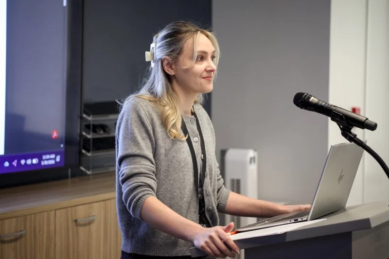 Young woman giving a presentation at a podium with a microphone, laptop, and a large screen behind her.