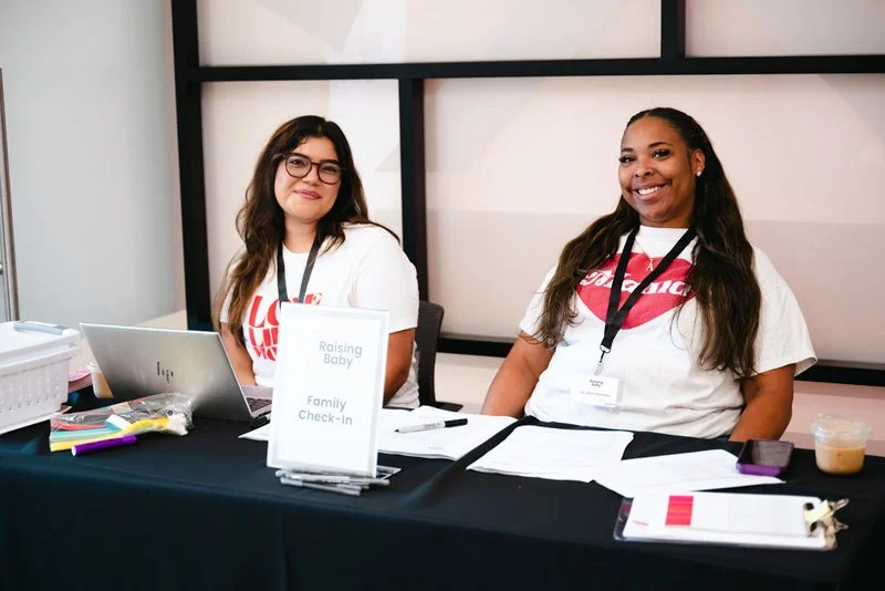 Two women sitting at a check-in table for a family event, smiling at the camera. The table has a sign that says "Raising Baby Family Check-In," with laptops, documents, and a drink on it.