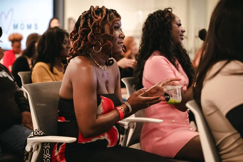 A woman with dreadlocks sitting in a conference room, holding a small container of food, wearing a strapless dress with floral design, surrounded by other women attending the event.