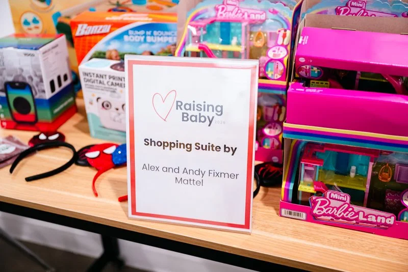 A display of children's toys including headbands, a Spider-Man mask, and Barbie Land playsets, with a sign reading 'Raising Baby 2026 Shopping Suite by Alex and Andy Fixmer Mattel'.