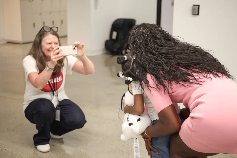 A woman with glasses on her head is crouching and taking a picture of a person with a large, curly-haired mask, who is holding a small stuffed dog. The woman appears to be smiling.