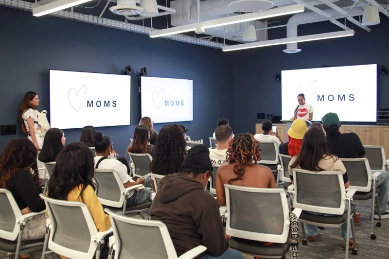 A woman is speaking at a presentation titled 'MOMS' in a room with seated audience members.
