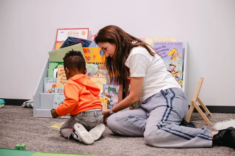 A woman and a young child sitting on a carpeted floor, looking at books together with a bookshelf filled with colorful children's books behind them.