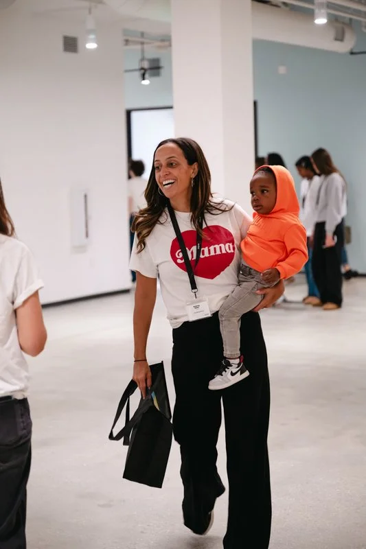 A woman smiling and holding a young child in an indoor space with white walls, other people in the background, and modern lighting.