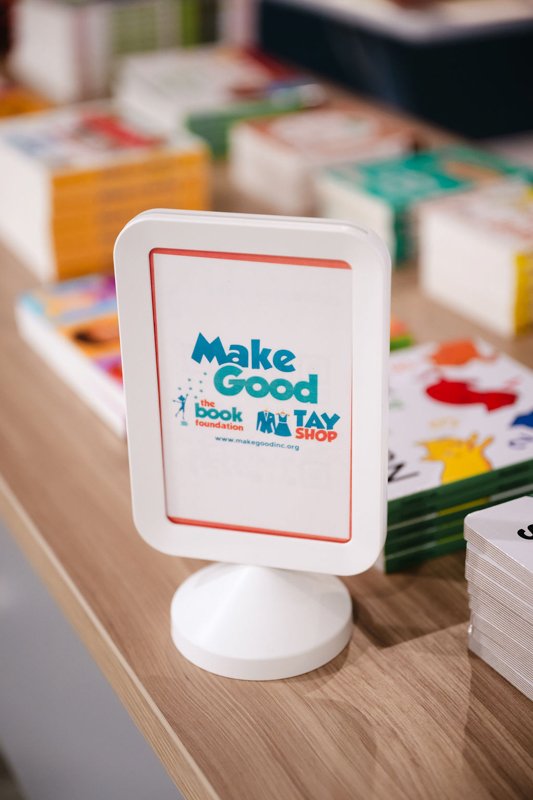 Sign on a table promoting the 'Make Good' book and toy store, with stacks of colorful books and games in the background.