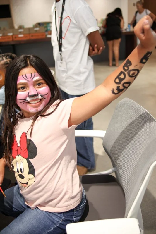 Girl with face paint resembling a cat, wearing a Minnie Mouse t-shirt, smiling and showing off black animal-print arm tattoos in a community or event space.