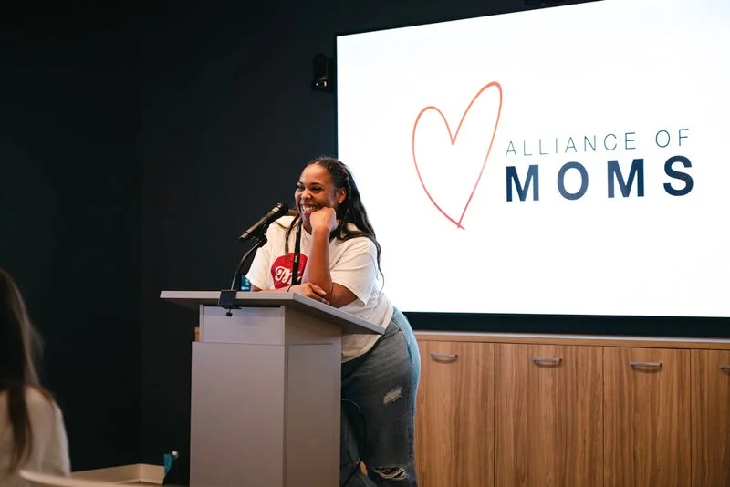 A woman standing at a podium, speaking into a microphone during an event for the Alliance of Moms, with a large screen behind her displaying the organization's logo.