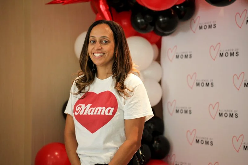 A woman smiling at an event, wearing a white T-shirt with a red heart and the word 'Mama' on it, standing in front of a backdrop with the word 'MOMS' and red and black balloons.