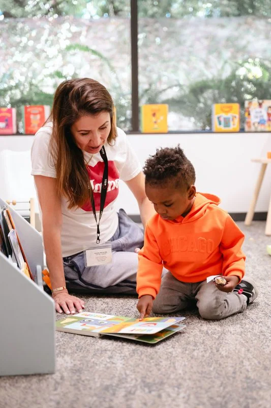 A woman and young boy reading a book together on the floor in a library or learning space.