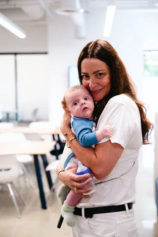 A woman with long brown hair holding a baby in an office setting.