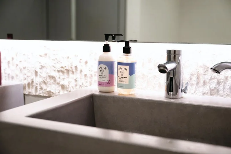 Two bottles of body lotion and body wash on a bathroom sink counter, with a mirror and textured wall background.