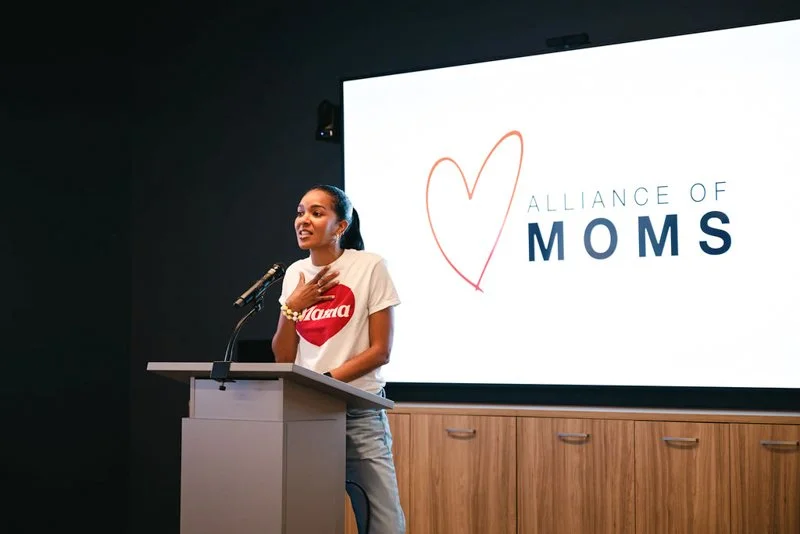 A woman at a podium speaking at an event with a large screen behind her that displays the logo 'Alliance of Moms' with a heart outline. The woman is wearing a white t-shirt with red print and has her hand on her chest.