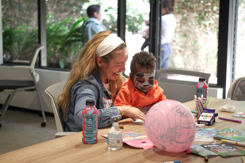 A woman with a white headband and long red hair, smiling, sits at a table with a young boy with face paint, engaged in arts and crafts. The table has a pink balloon, bottles of water, markers, and coloring supplies. Large windows in the background re