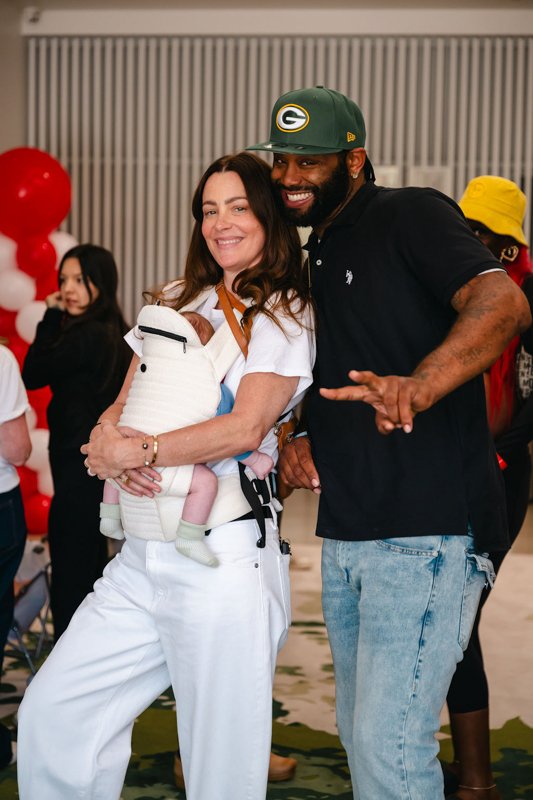 A woman holding a baby and a man standing next to her at a social event with red balloons in the background.