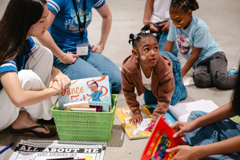 A group of children and adults sitting on the floor, engaging in a storytelling or learning activity, with some books and colorful materials around.