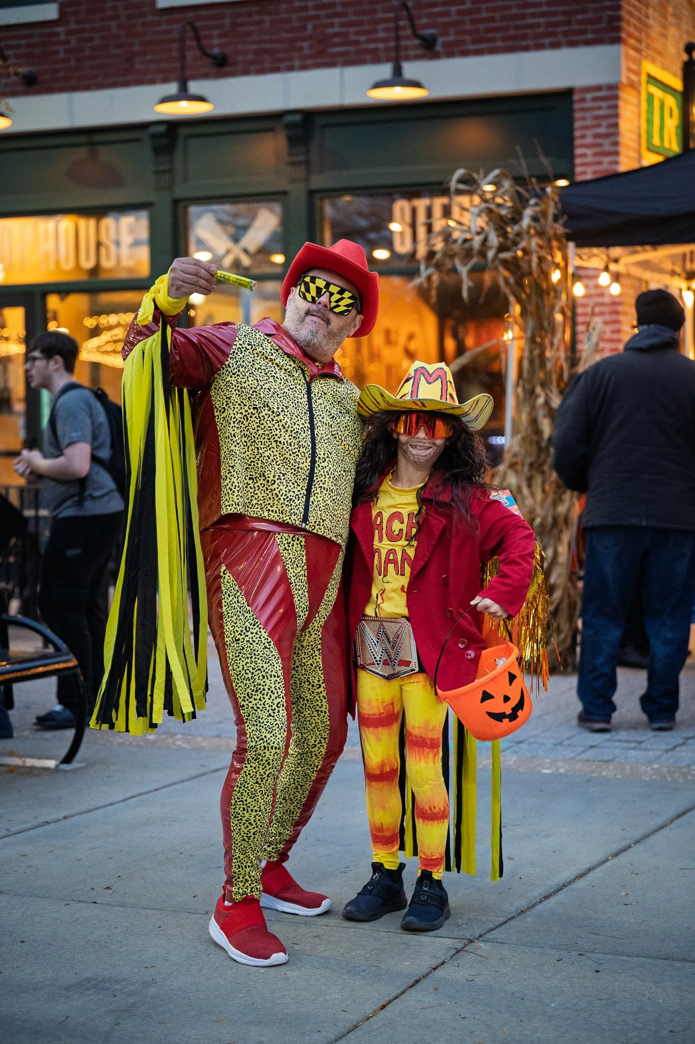 Two people dressed in colorful, flamboyant costumes posing for a photo outside during Halloween. The man is wearing a red hat, yellow and red jacket, matching pants, red shoes, and yellow sunglasses with black patterns. The girl is wearing a decorate