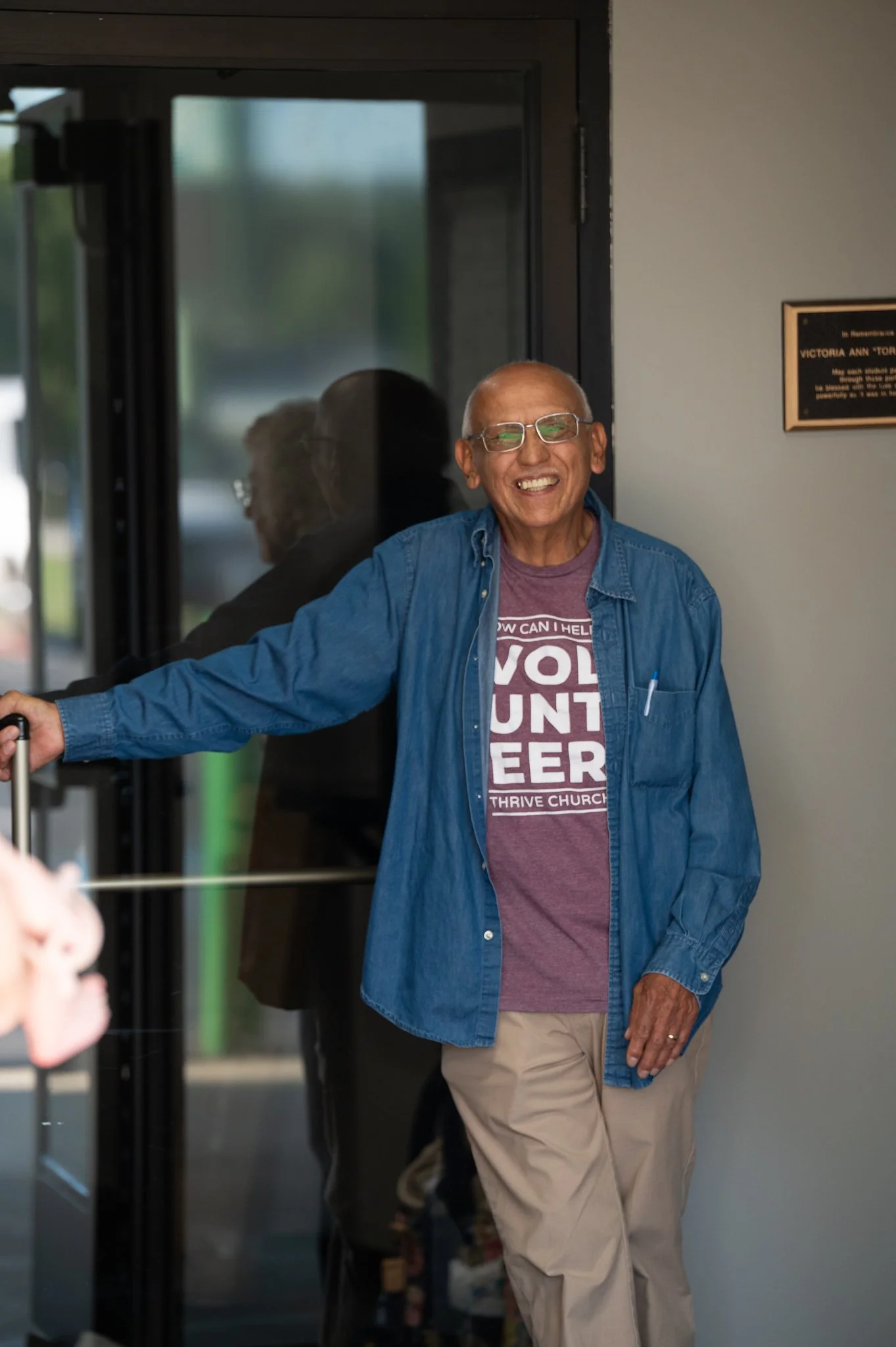 An elderly man with glasses, smiling and wearing a denim shirt over a maroon t-shirt, enters a building through a glass door.