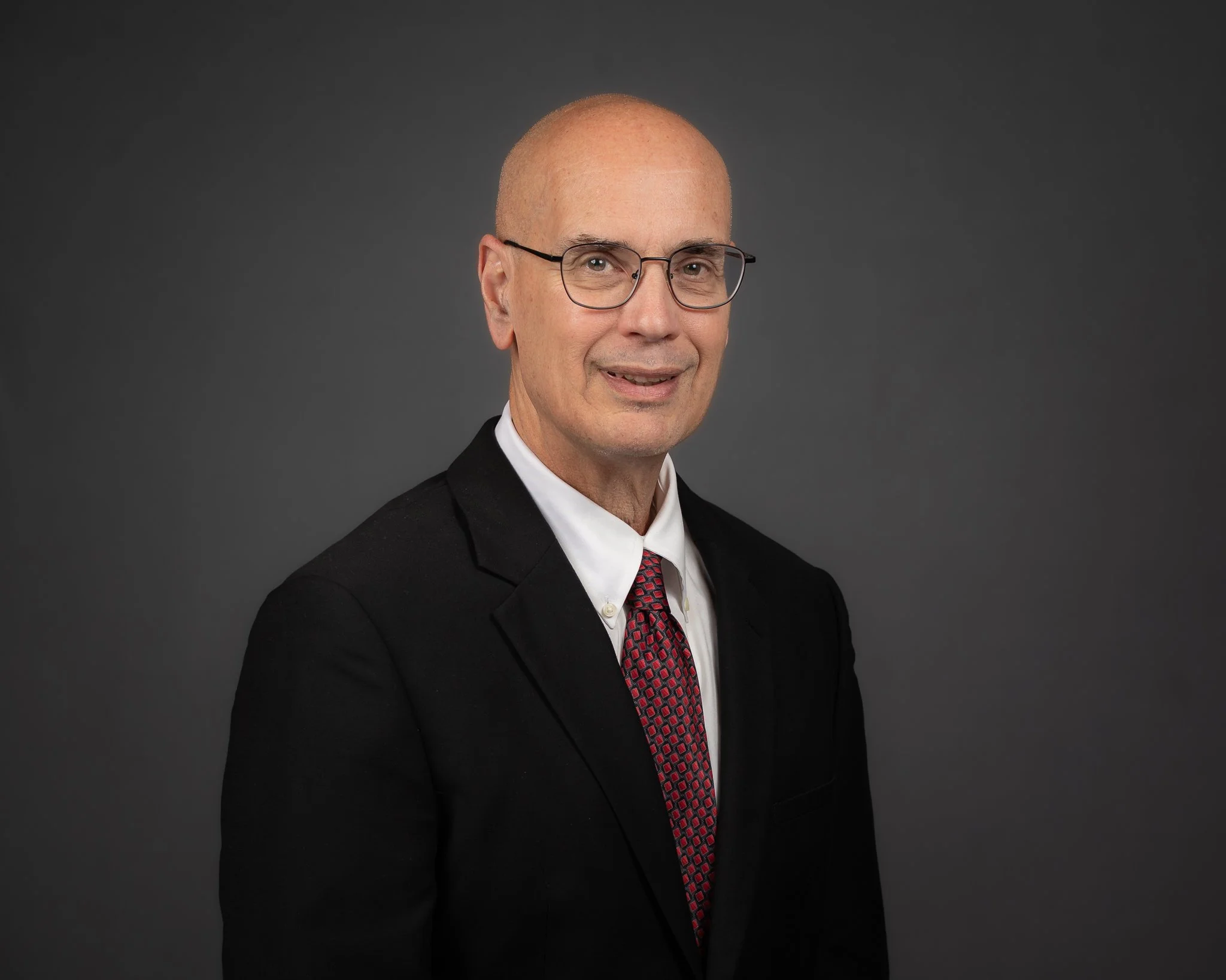 A bald man wearing glasses, a black suit, white shirt, and red patterned tie, standing against a dark gray background.