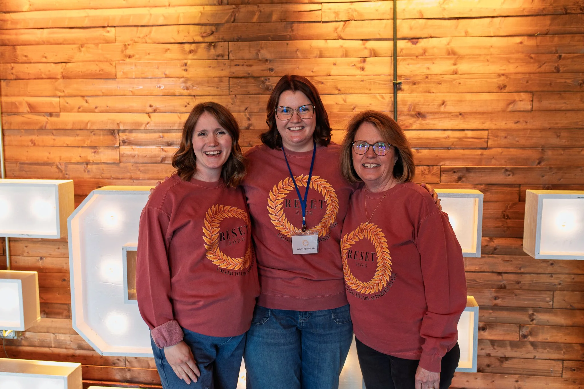 Three women standing close together, smiling, in front of a wood-paneled wall. They are wearing matching maroon shirts with a circular logo that says "RESET 2026" and a laurel wreath design. One woman has a name tag that reads "Leigh Flegge-Schlei".