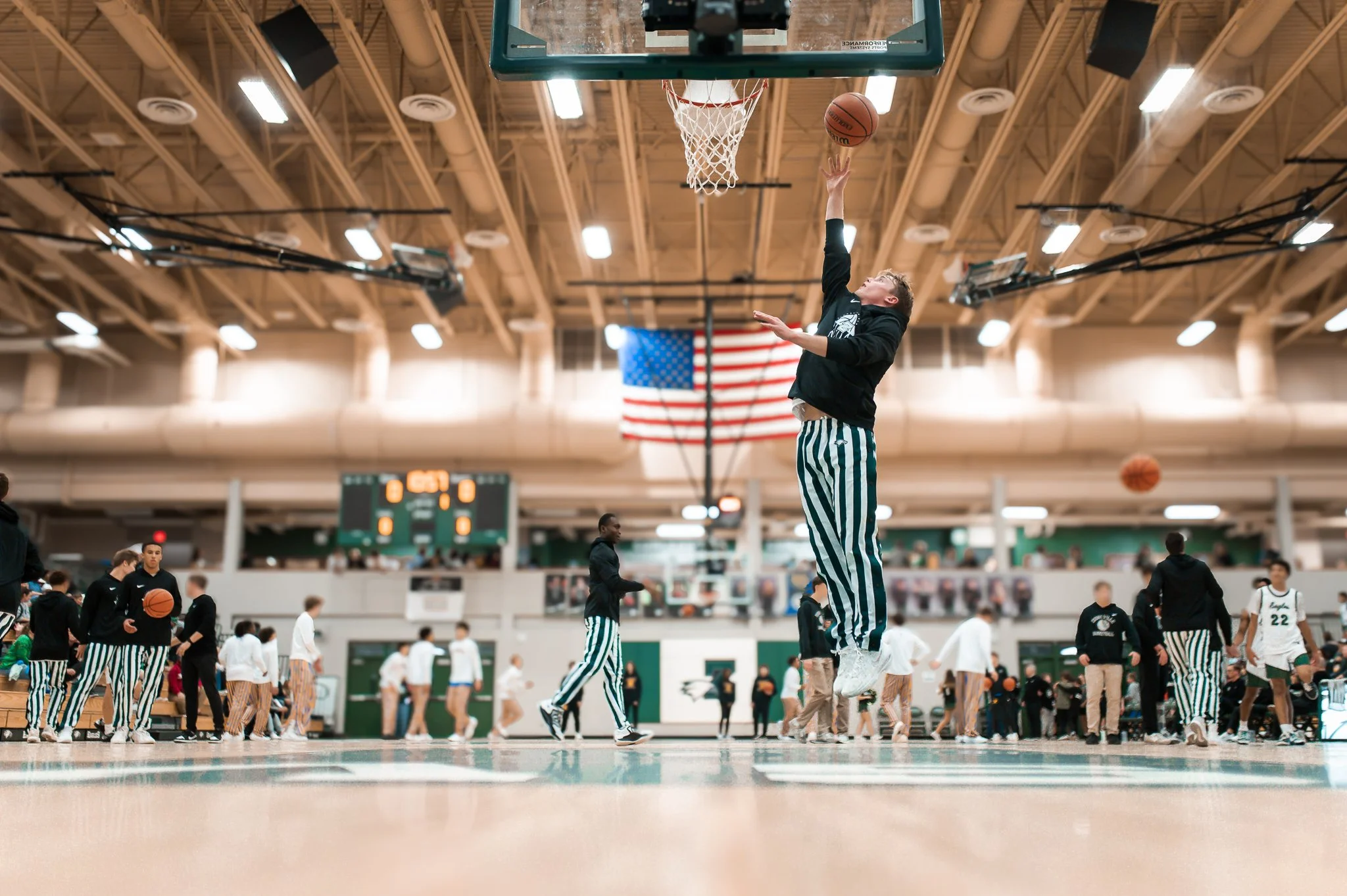A young man wearing striped pants and a black long-sleeve shirt is jumping to make a basketball shot at a basketball hoop in an indoor gymnasium. Other players are visible in the background, and an American flag hangs above the court.