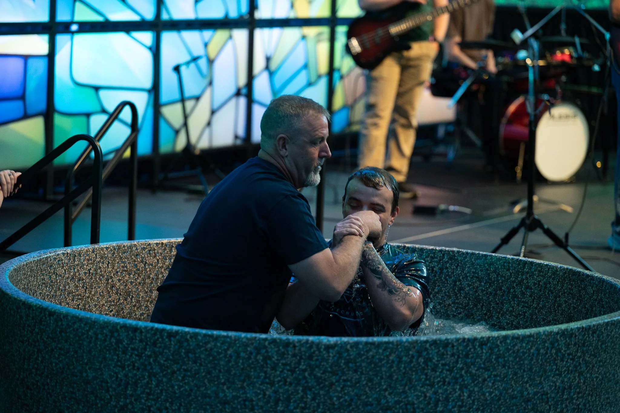 A man with a beard is baptizing a person in a small, round pool, in front of a colorful, geometric stained glass window. A musician is seen in the background near musical equipment.