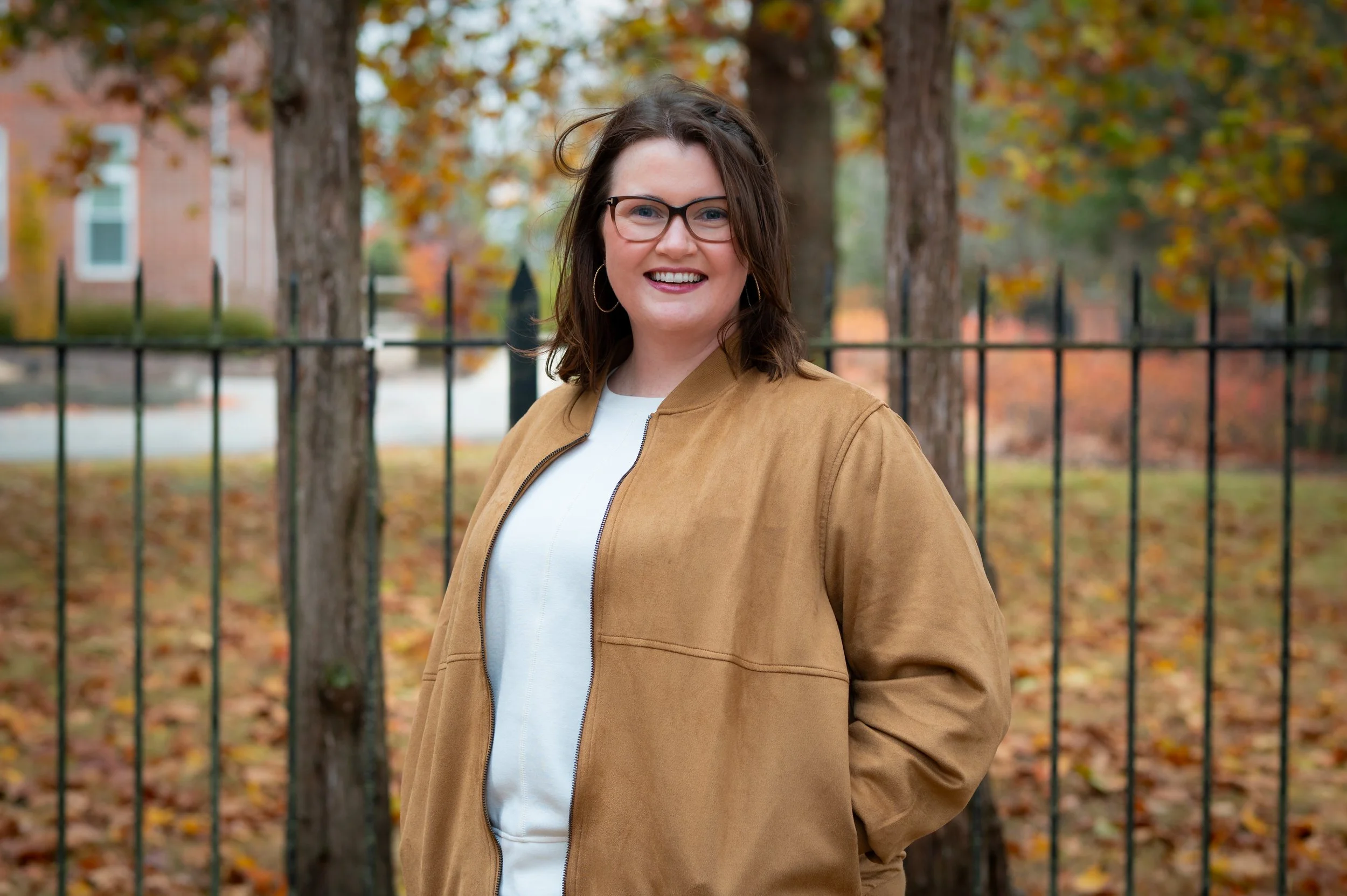 A smiling woman with glasses and hoop earrings standing outdoors in fall, wearing a tan jacket and white shirt, with trees and leaves in the background.