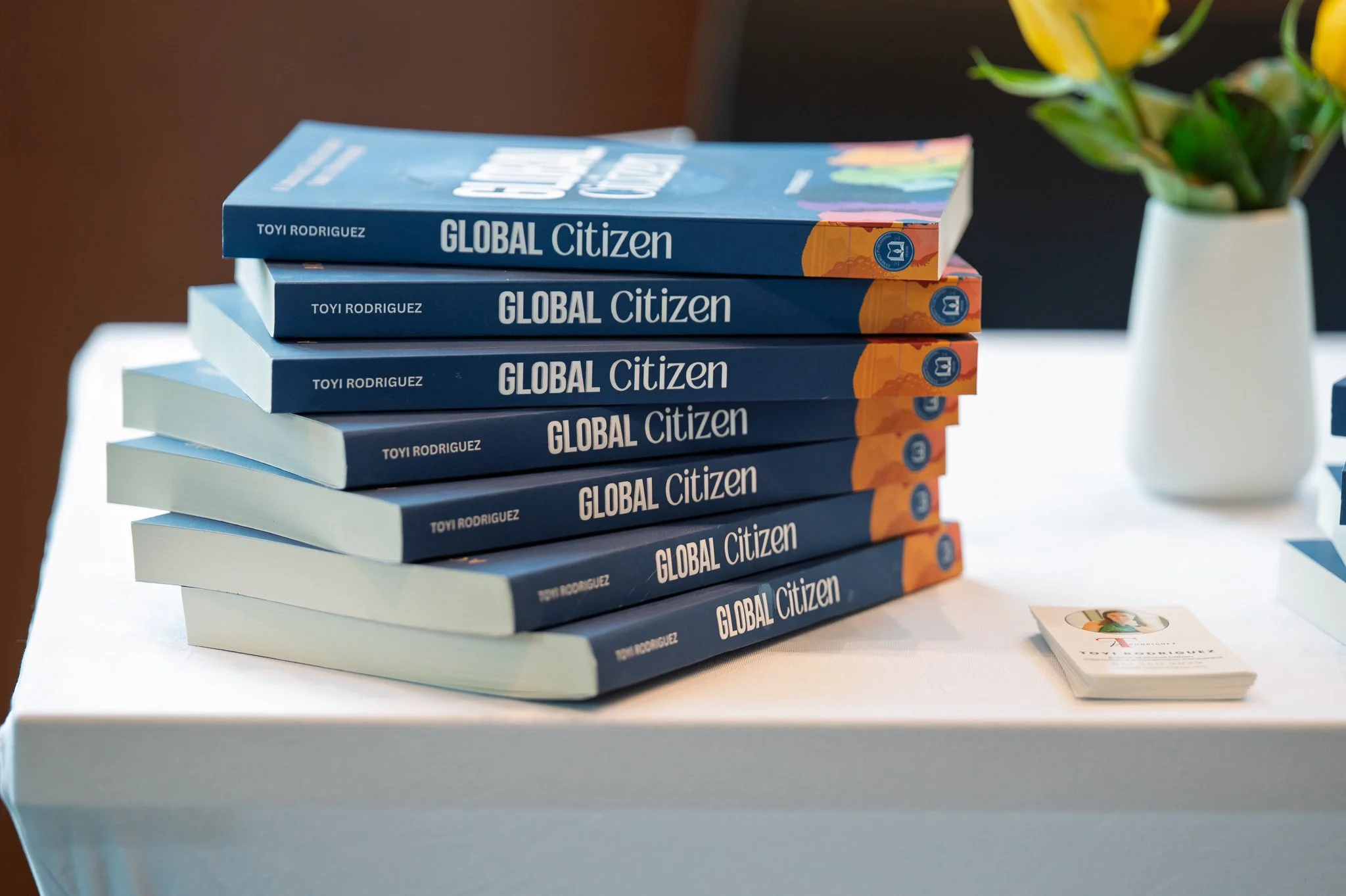 Copies of the book Global Citizen sit on a table next to business cards and a vase of yellow flowers.