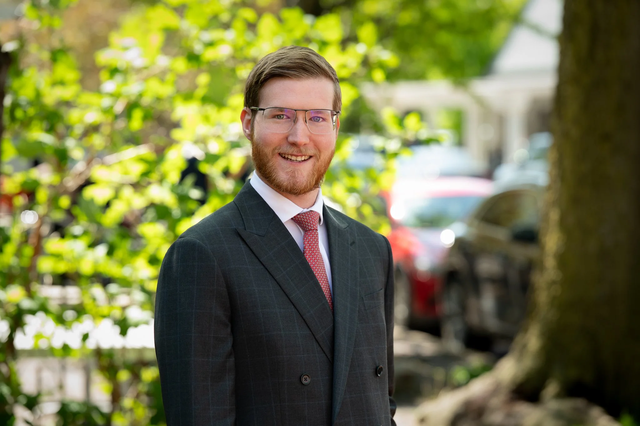 Young adult male wearing glasses, a black suit, and white dress shirt with red tie, poses for an outdoor headshot with trees and cars in the background.