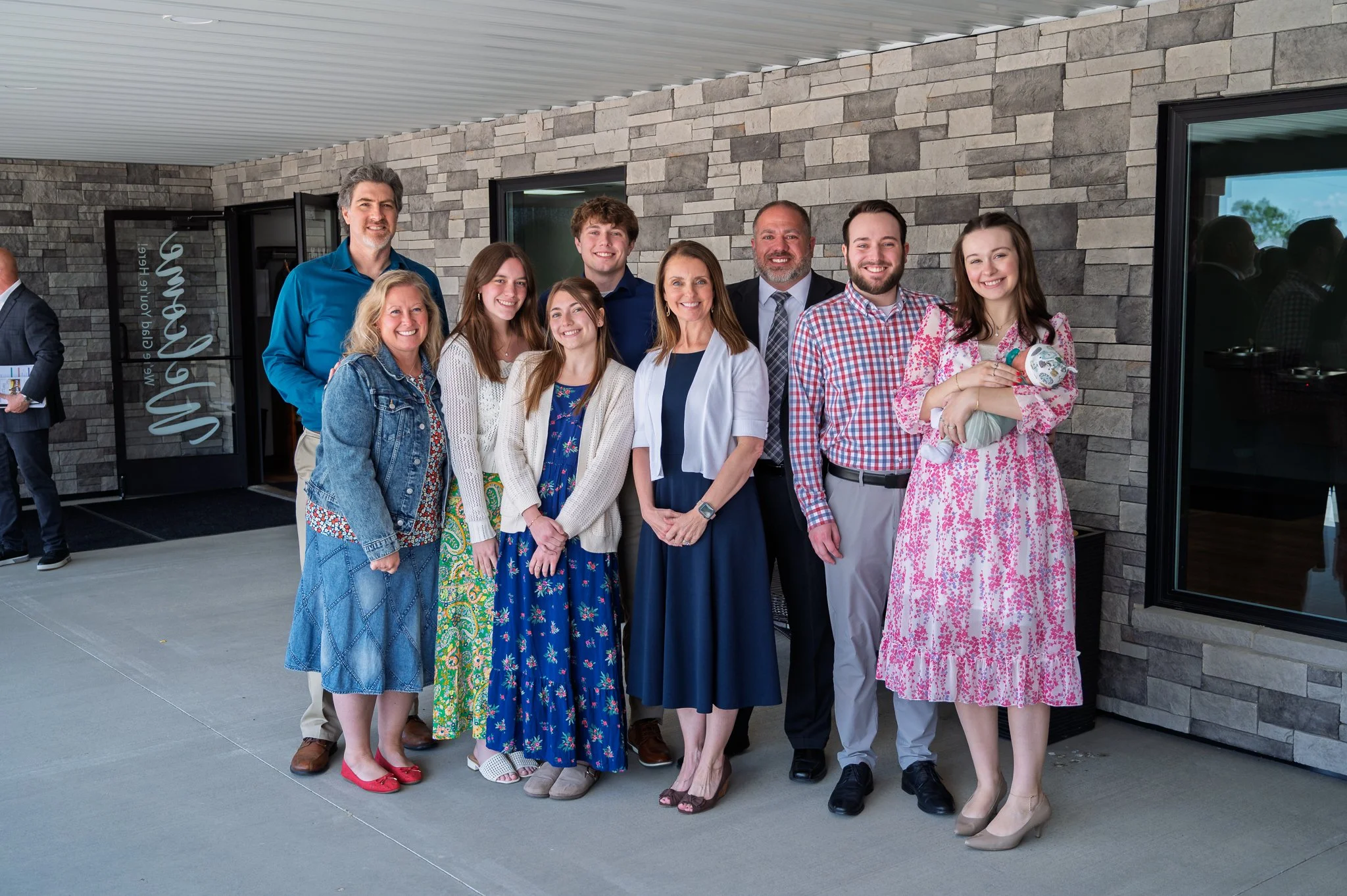 A group of eleven people standing outside a building, smiling at the camera during daytime. The background features a brick wall and windows, with some reflections visible. The group includes men and women of various ages, dressed in casual and semi-