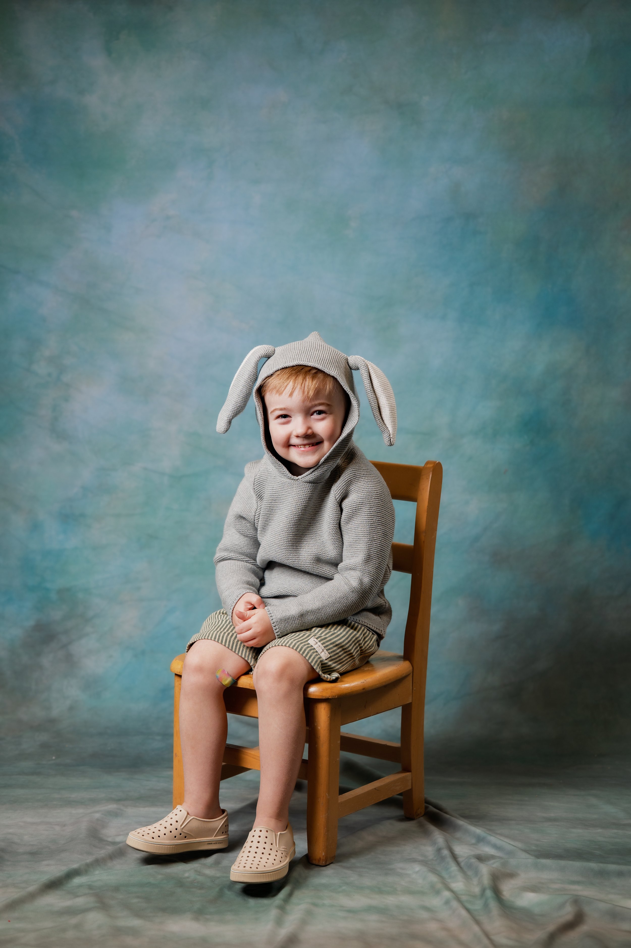 A young boy in a hooded sweater with rabbit ears sits in a wooden chair posing in front of a blue green background.