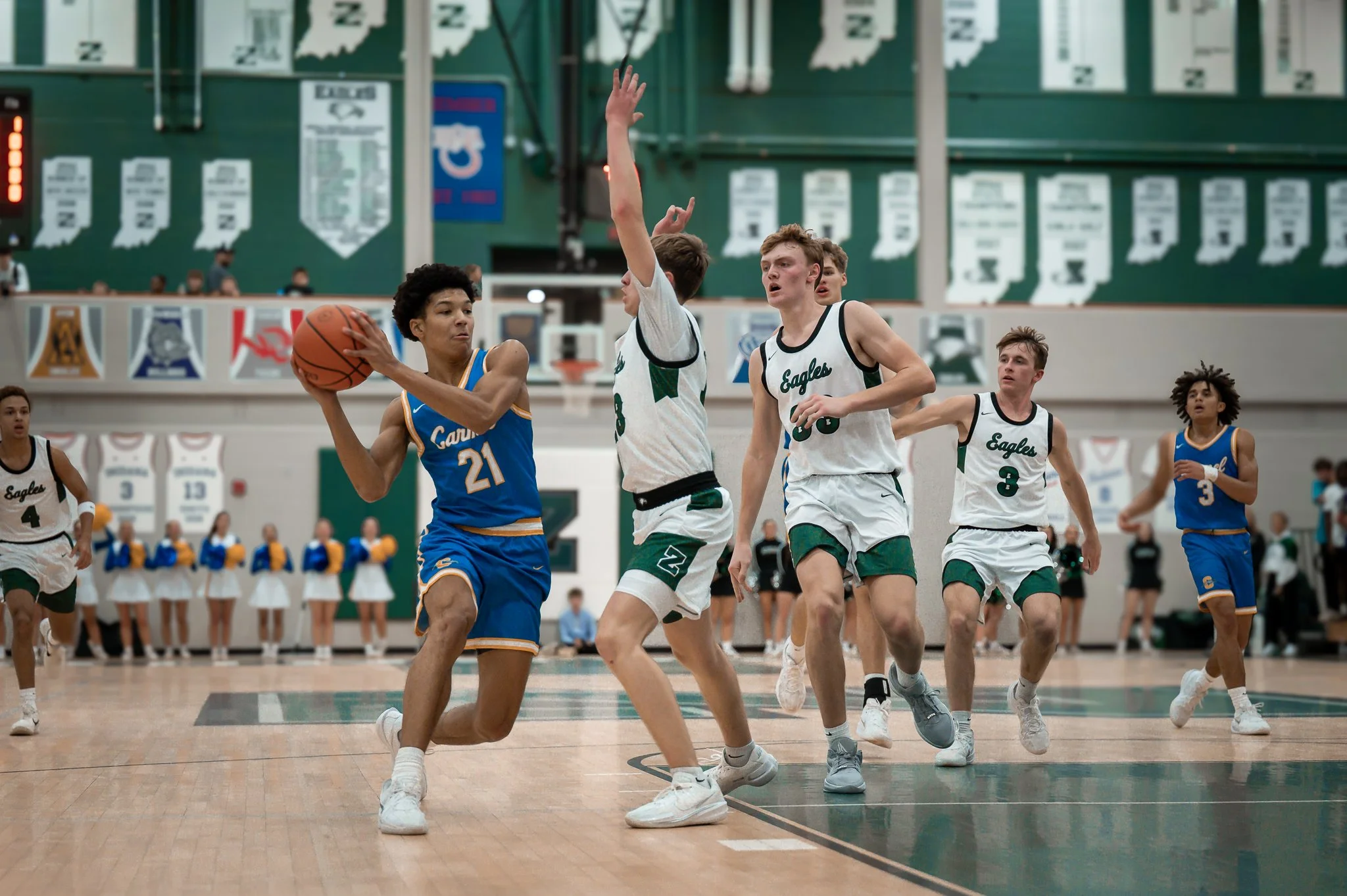 A basketball game with players in white and blue jerseys inside a gymnasium. One player in blue is holding the ball, while several players in white are defending.