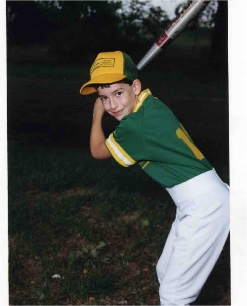 Little leaguer poses for a photo while holding a bat wearing a gold and green uniform and hat with white baseball pants.