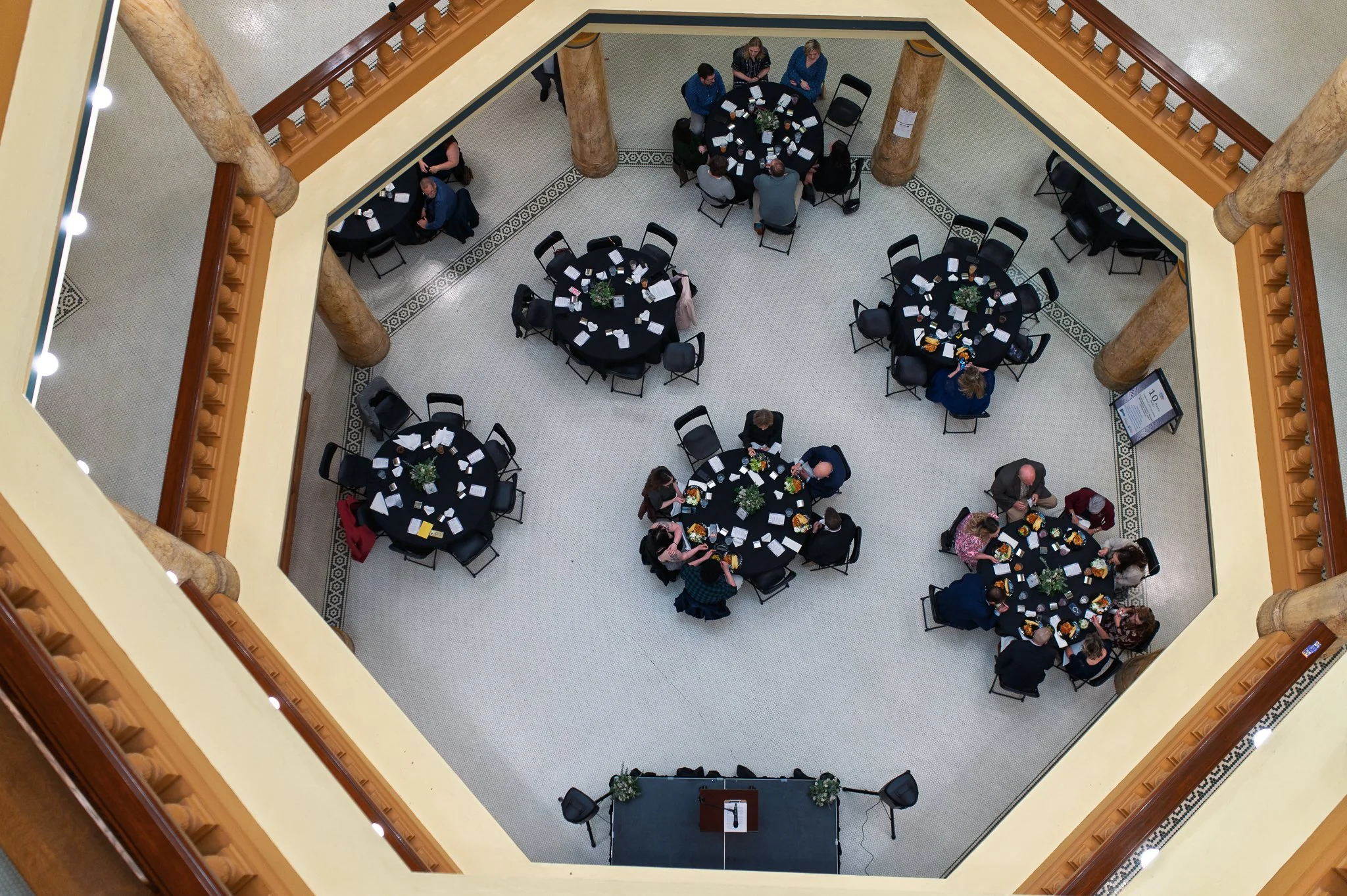 View from above of a conference or banquet room with round tables set for an event, surrounded by people and floral centerpieces, with a lectern at the front.
