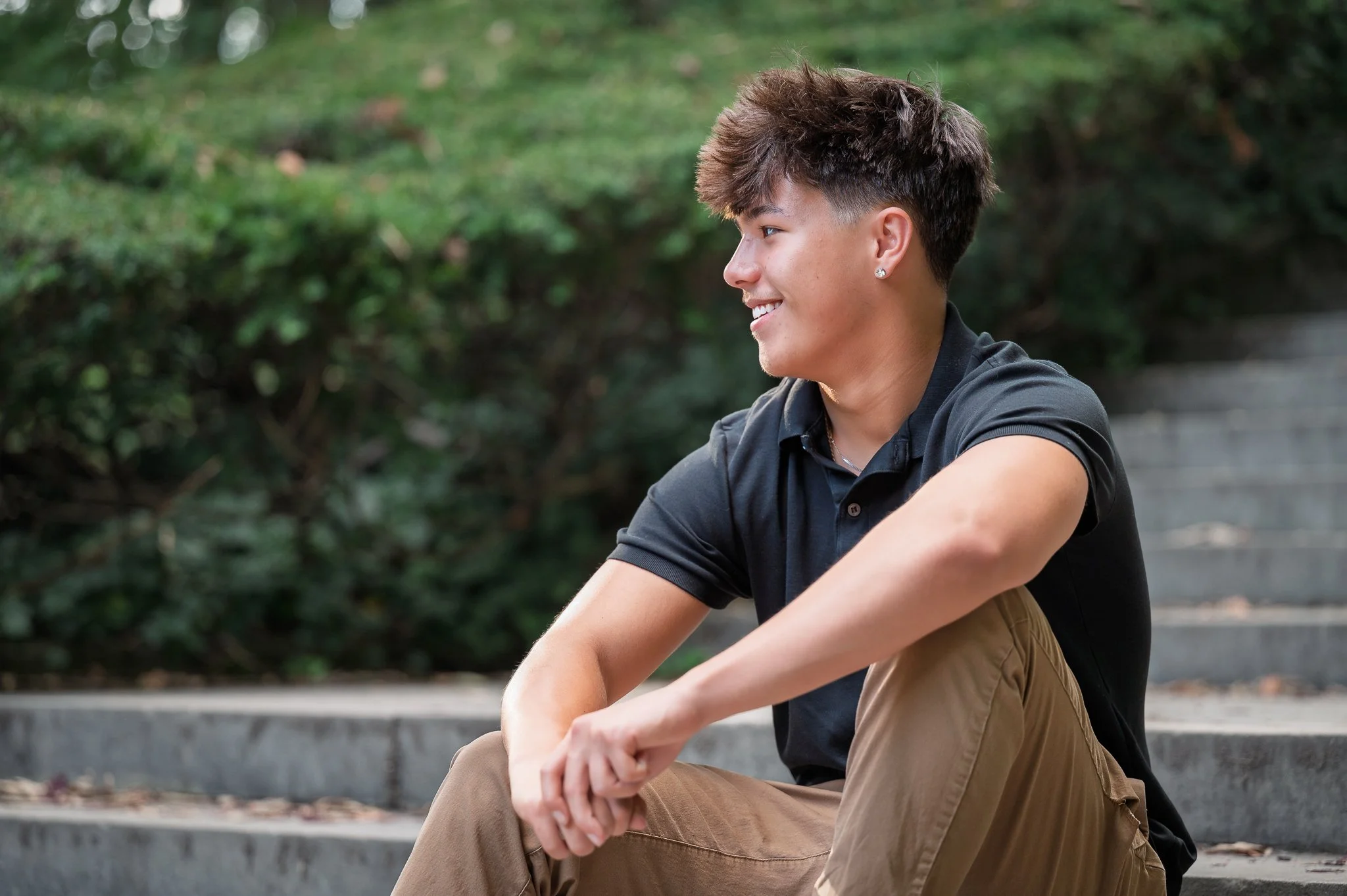 A young man sitting on outdoor steps, smiling and looking to his right, with green bushes behind him.