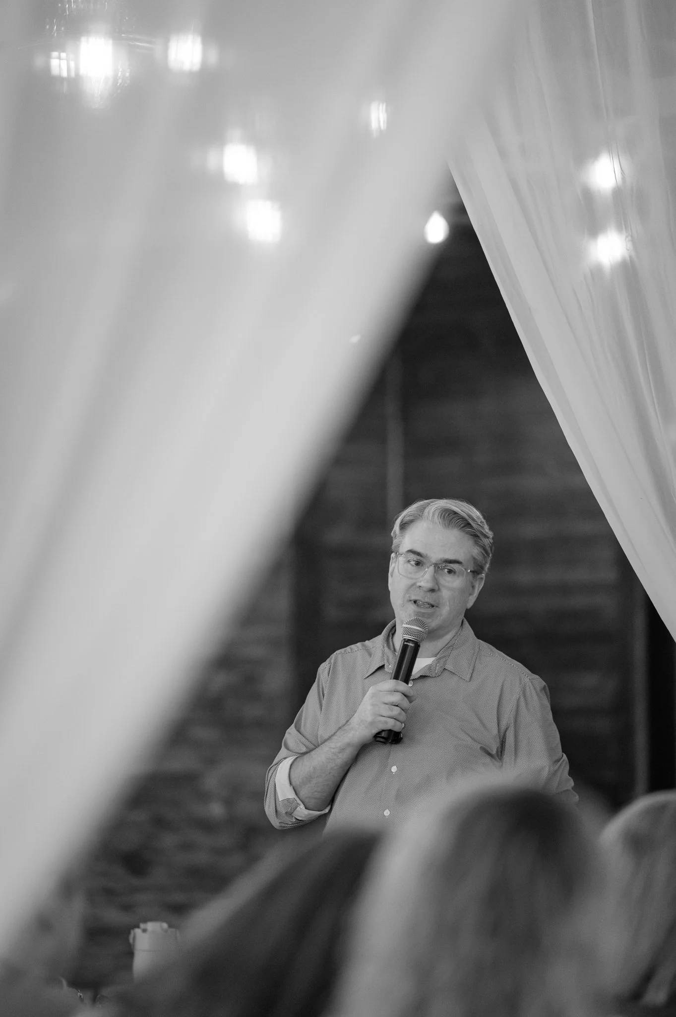 A man with glasses and gray hair, wearing a gray button-up shirt, is speaking into a microphone. He appears to be giving a speech or presentation in a setting with dark wooden stairs in the background.