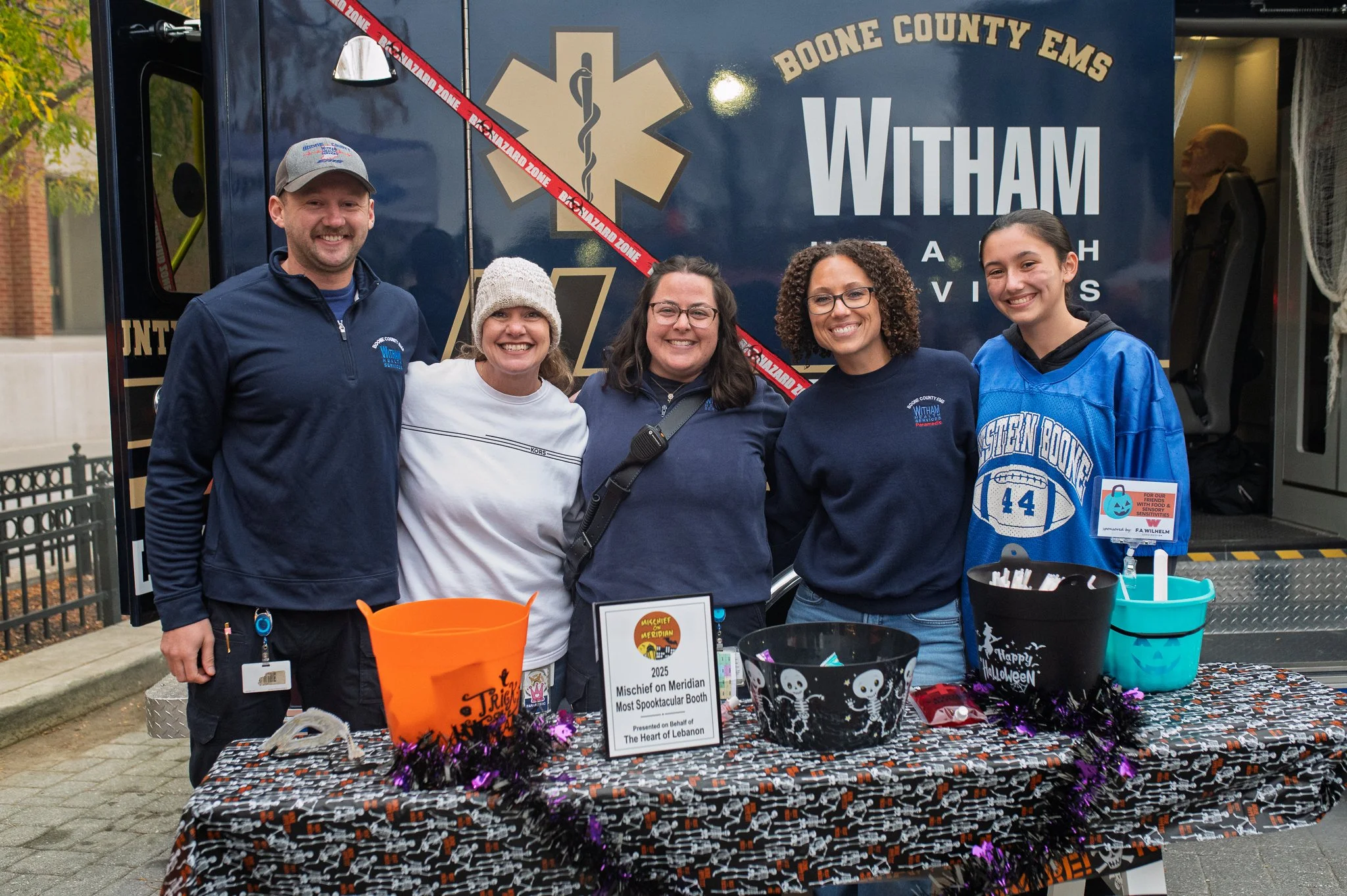 Group of five people smiling and standing behind a Halloween-themed table display outdoors, with a decorated truck in the background. The table is decorated with Halloween decorations, including buckets and a sign. The sign indicates they are partici