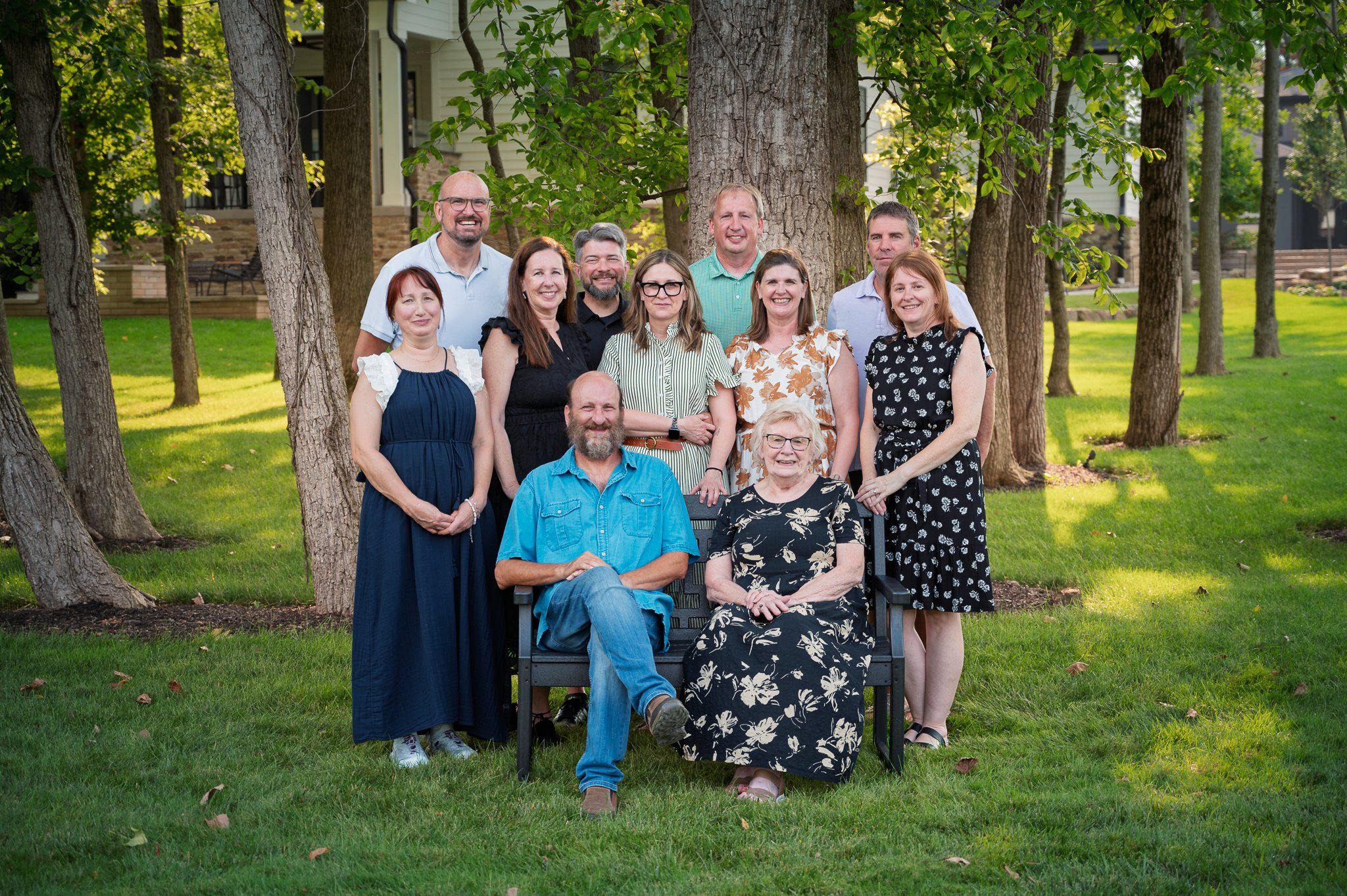 Group of eleven people posing outdoors in front of trees and a house, with some seated and others standing.