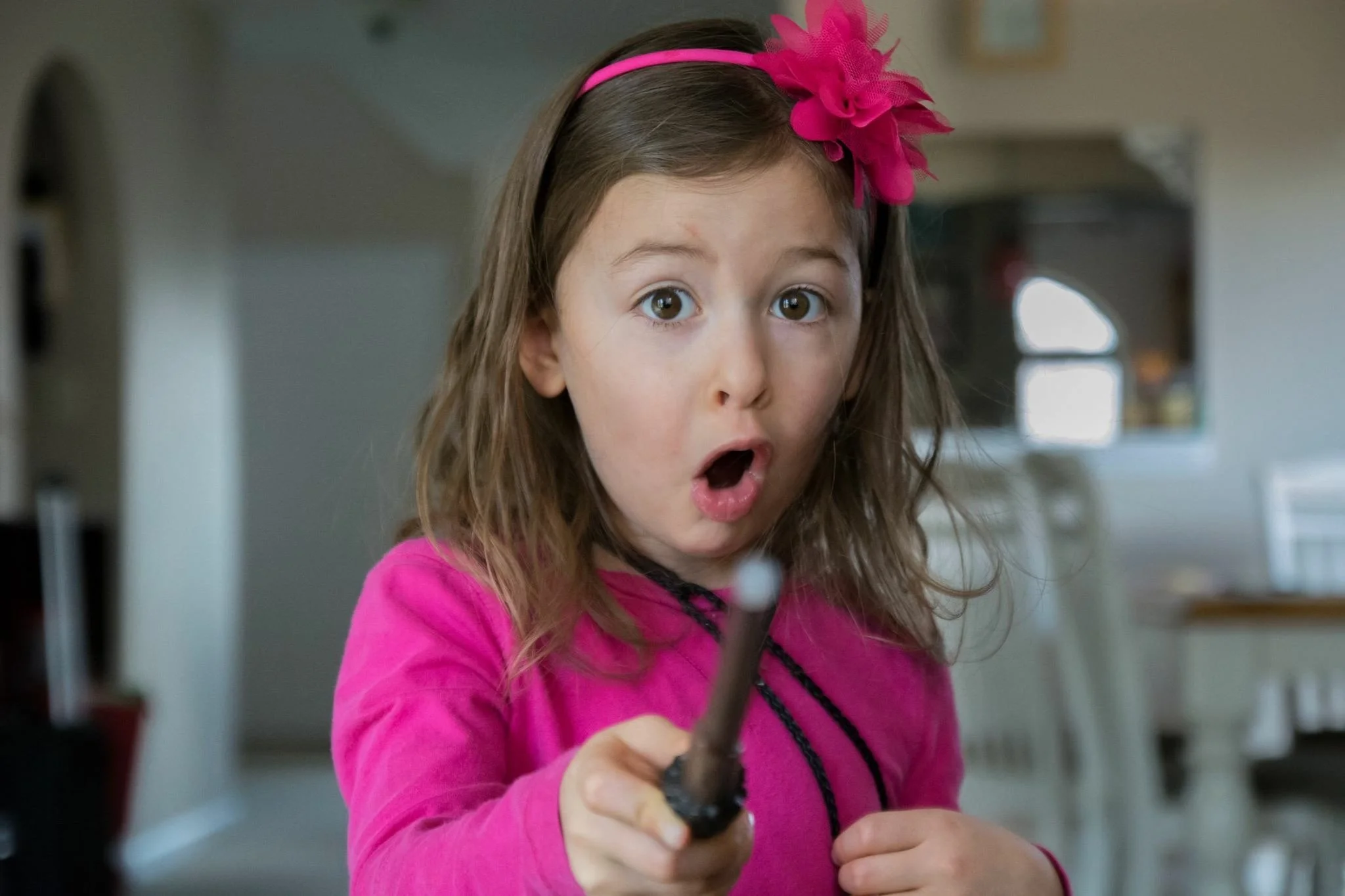 A young girl with long brown hair wearing a pink long-sleeve shirt and a pink headband with a large fluffy pink flower, holding a marker and looking surprised or shocked.