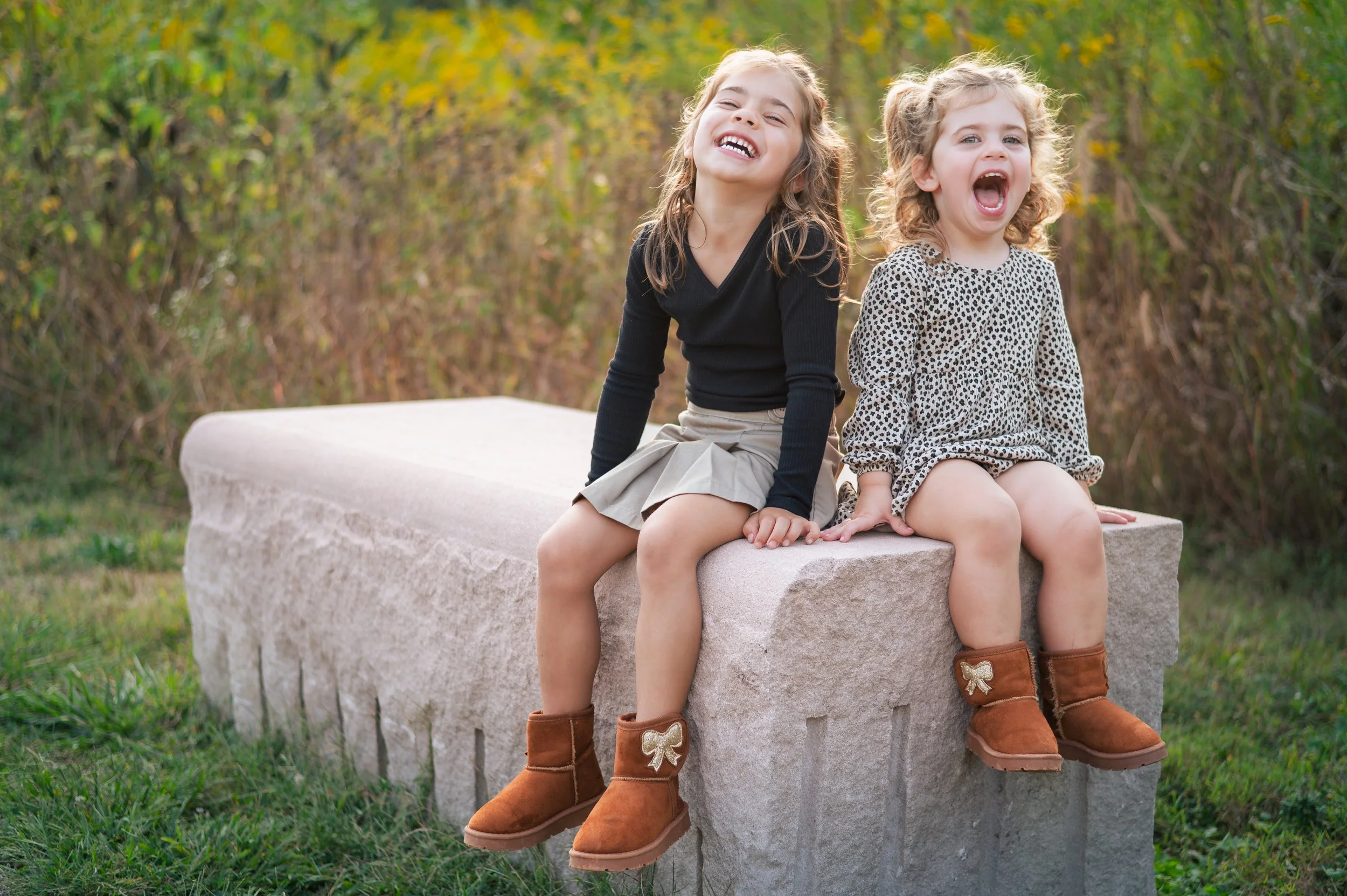 Two young girls sitting on a large stone bench outdoors, smiling and laughing, surrounded by green and yellow foliage.