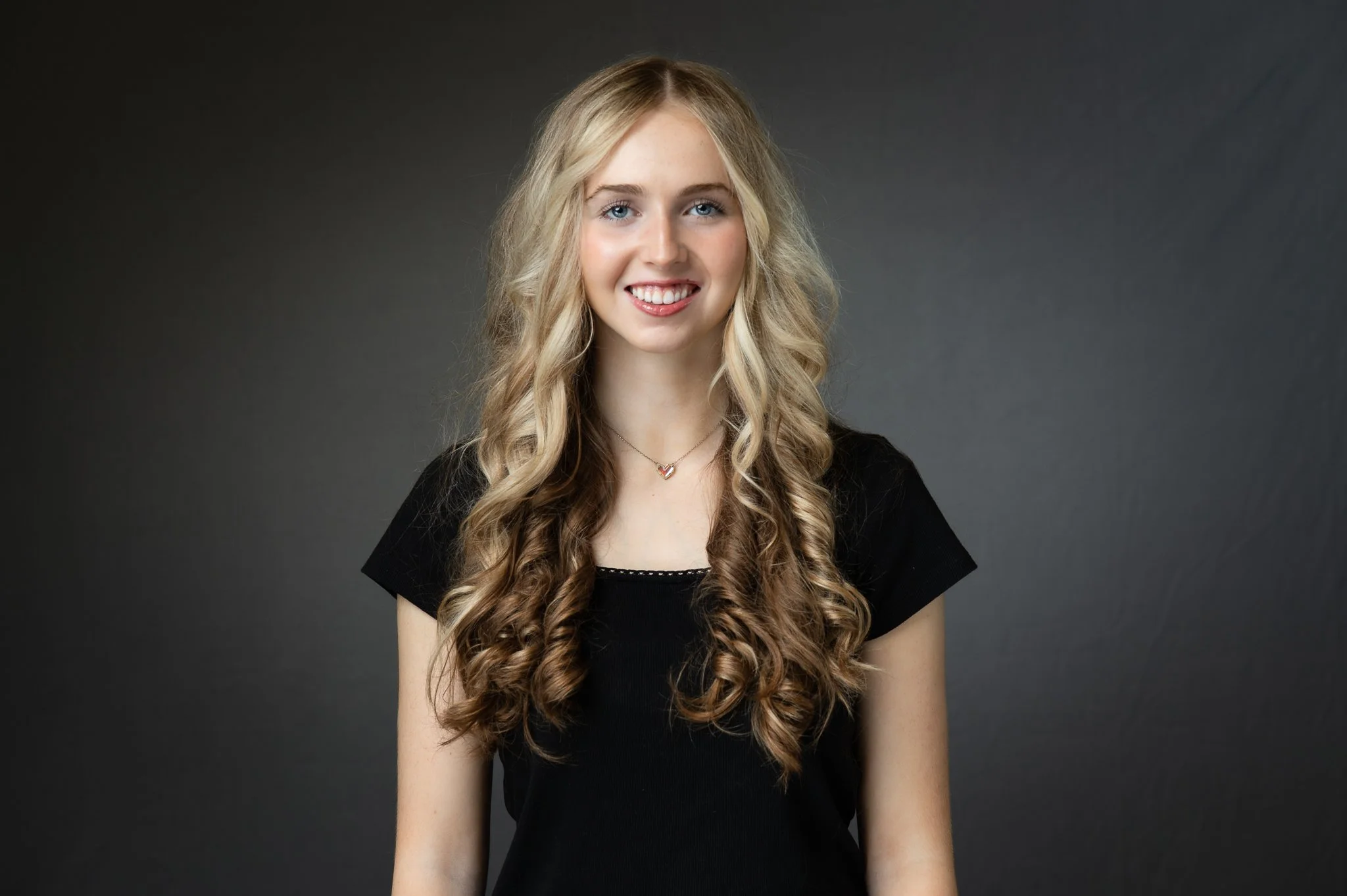 Portrait of a young woman with long, curly blonde hair wearing a black shirt and a heart-shaped pendant necklace, smiling against a dark gray background.