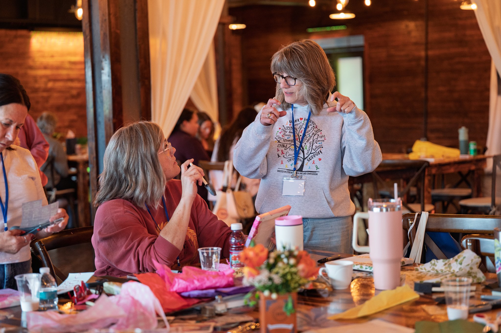 A group of women sitting around a table during a craft or social event, with one woman standing and engaging in conversation.