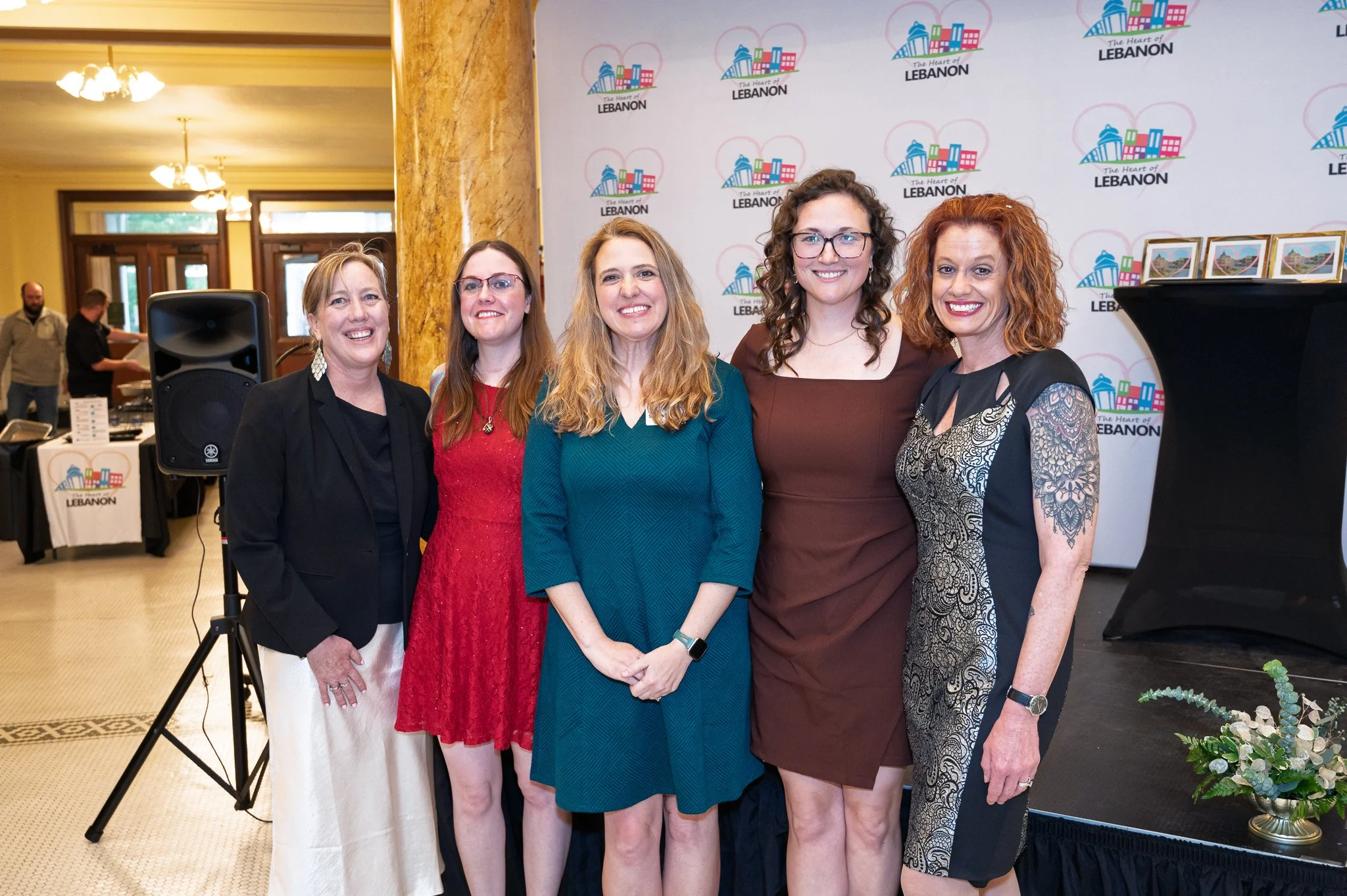 Five women standing together, smiling, at an event with a backdrop that reads 'The Heart of LEBANON'. They are dressed in business or semi-formal attire, and the background includes framed photos and event signage.