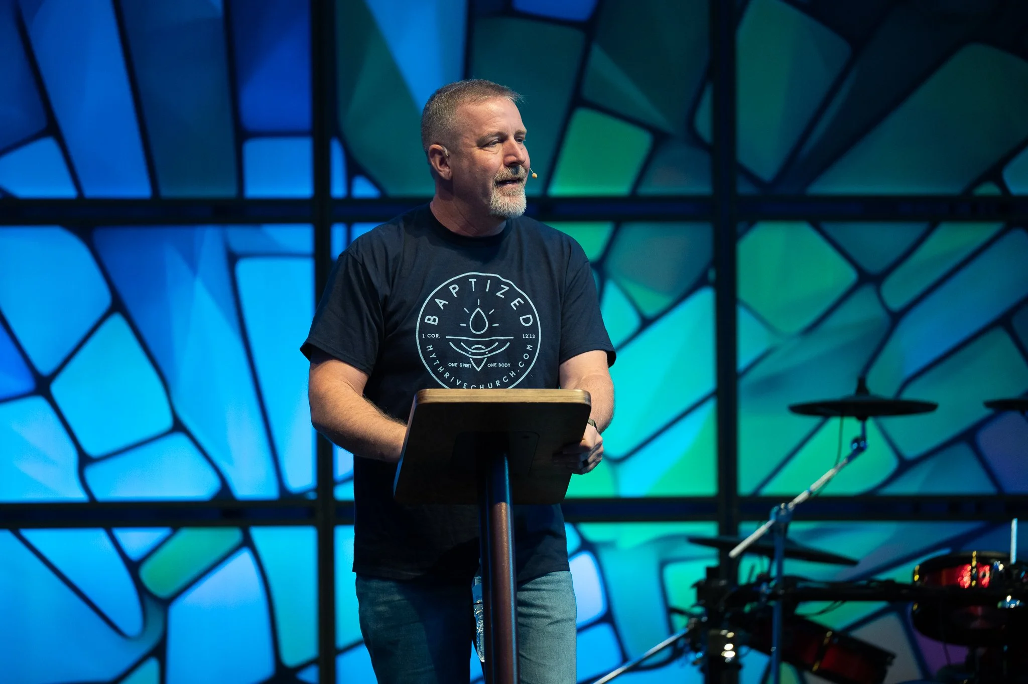 A man with short hair, a beard, and wearing a microphone headset stands behind a podium, speaking at an event with a colorful abstract background.