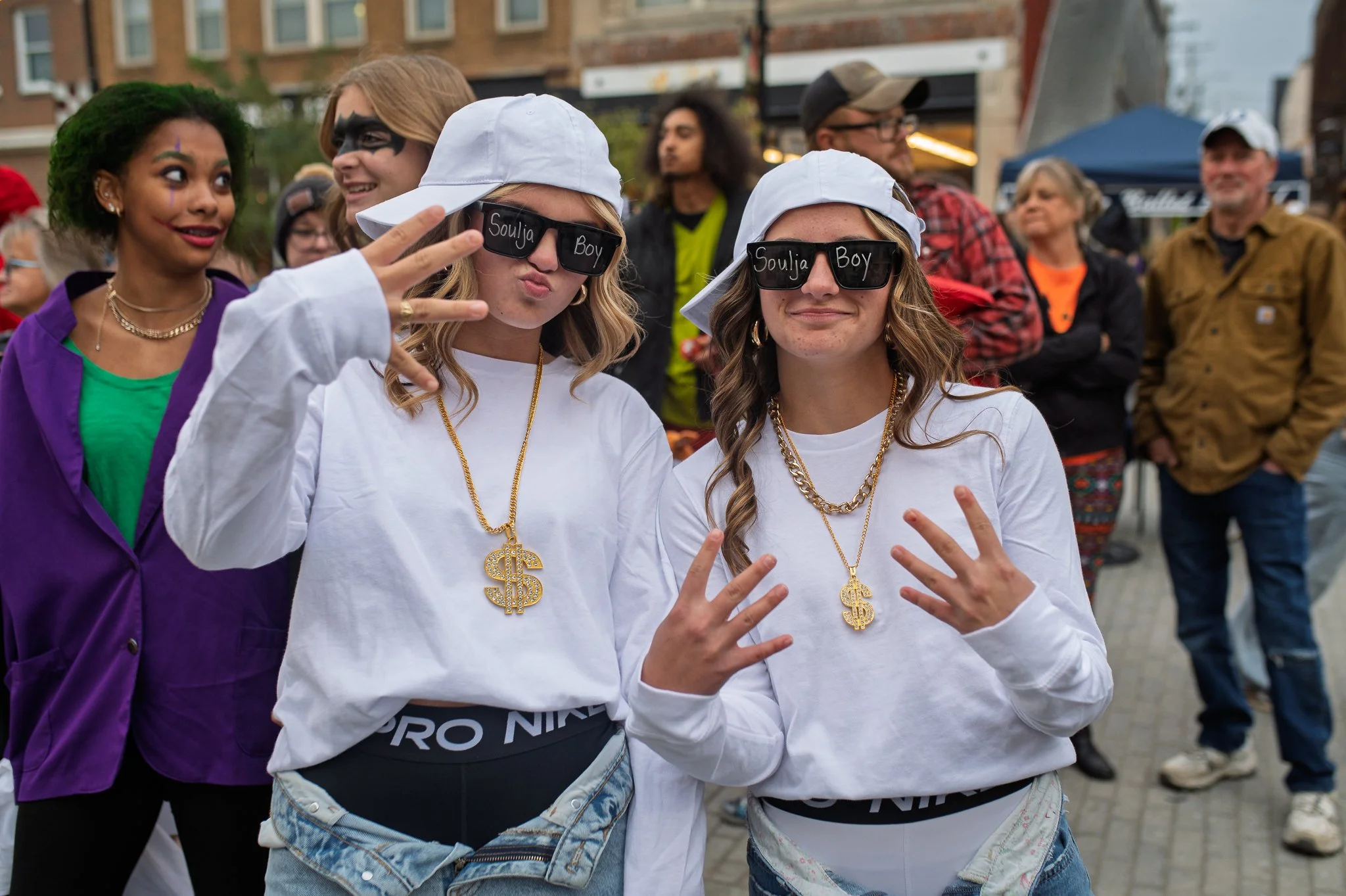 Two young women with sunglasses that say 'Soulja Boy,' wearing white caps backward, gold chains with dollar signs, and making peace signs, at an outdoor event with people in the background.