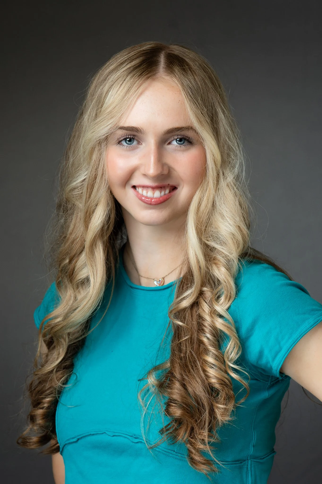 Portrait of a young woman with long, wavy blonde hair wearing a turquoise top, smiling with teeth showing, against a dark gray background.