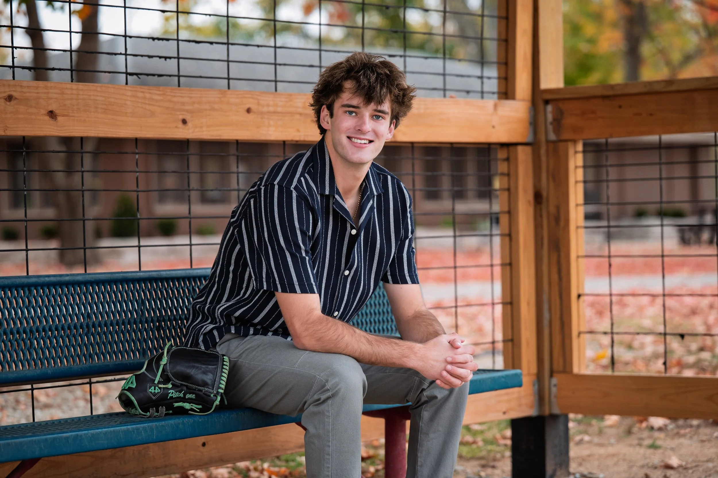 Young man sitting on a blue park bench, wearing a black and white striped shirt and gray pants, smiling, with a baseball glove placed on his lap, surrounded by a wooden fence with trees in the background.