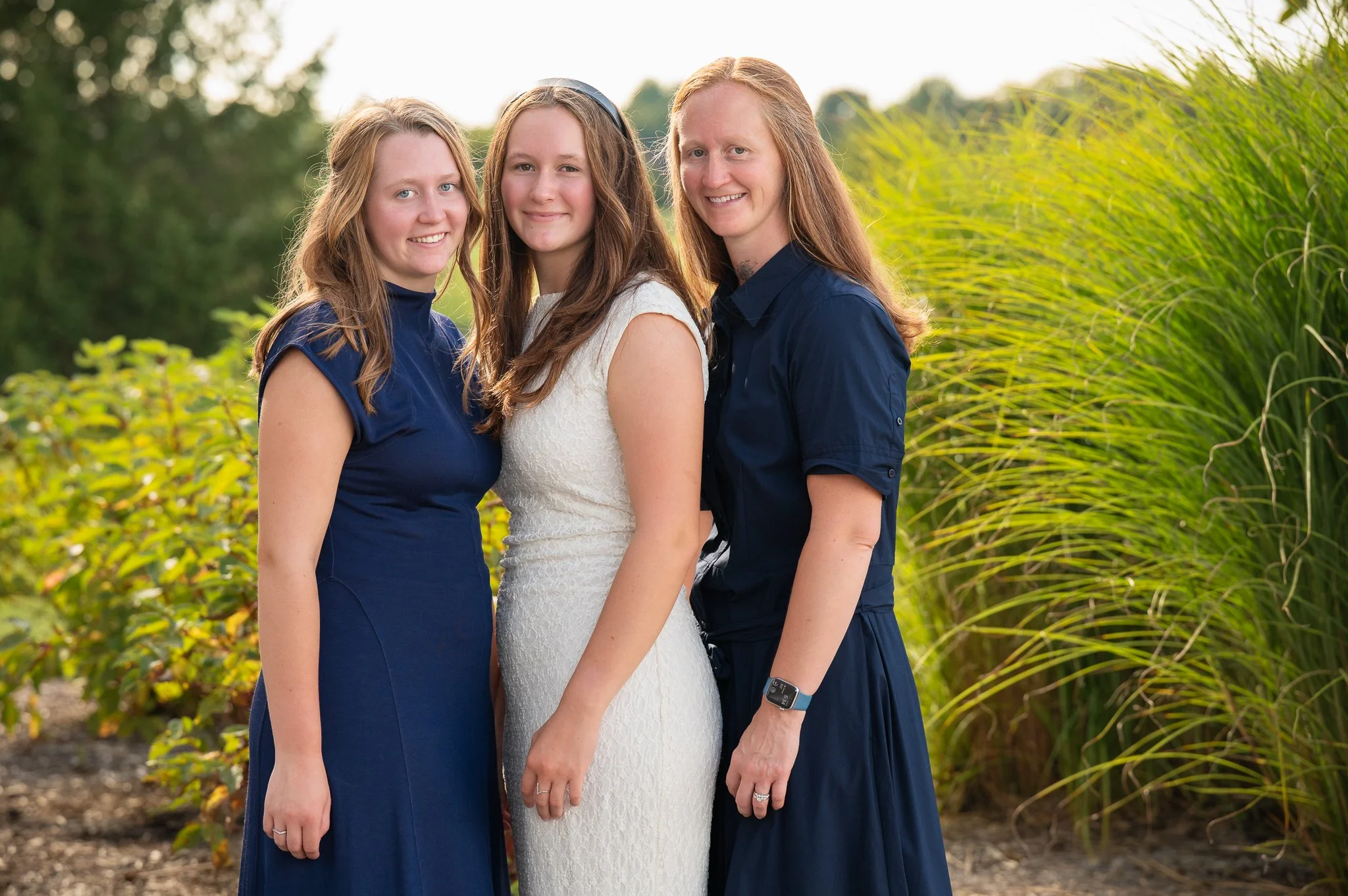 Three women standing outdoors in front of green plants, smiling and close together. One woman in a white dress, flanked by two women in dark blue dresses.