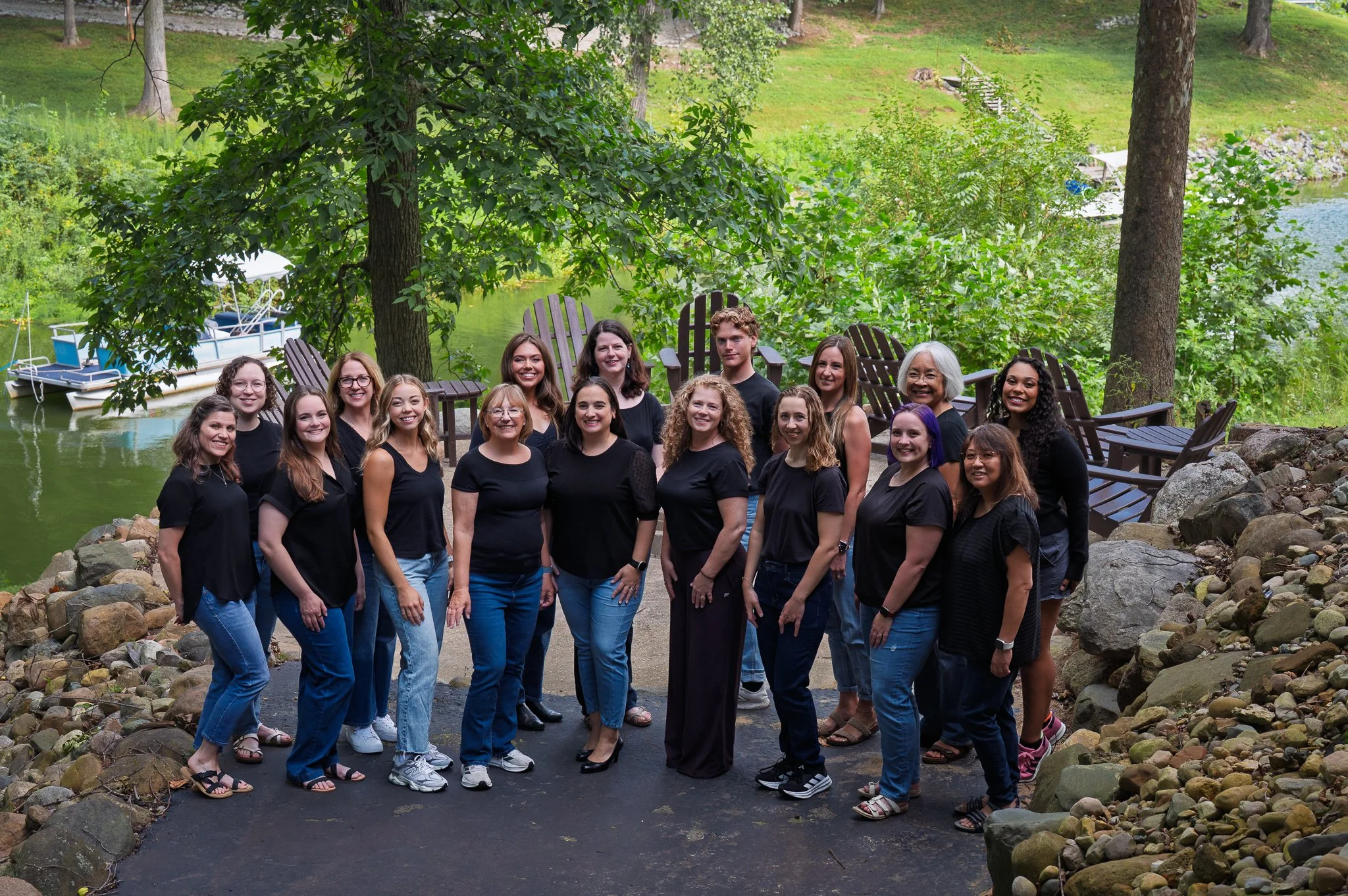 A group of fifteen women and one young man standing outdoors near a body of water with boats, surrounded by trees and rocks, smiling for a photo.
